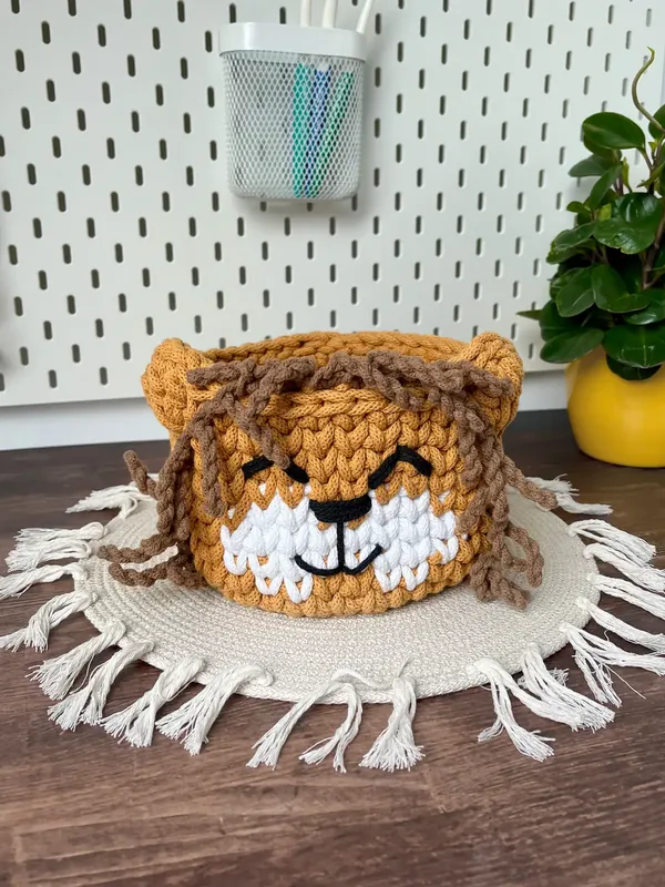 A mustard crochet lion basket with a smiling face and a loose brown mane, on a fringed mat