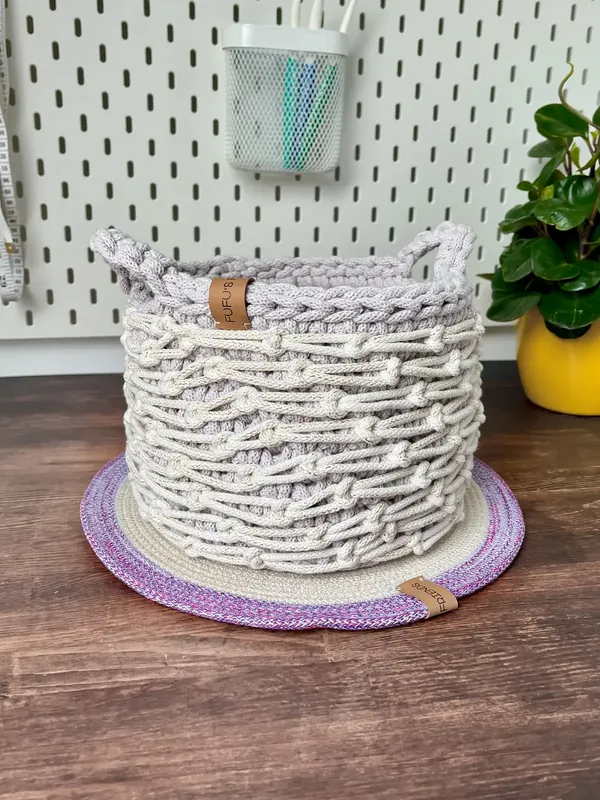 A cream knotted crochet basket with a lilac rim and two handles, on a lilac-edged mat